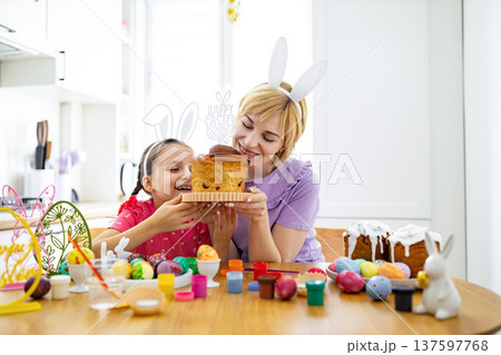 A mother and daughter wearing bunny ears excitedly sniff a traditional Easter cake while surrounded by painted eggs and baking supplies 137597768