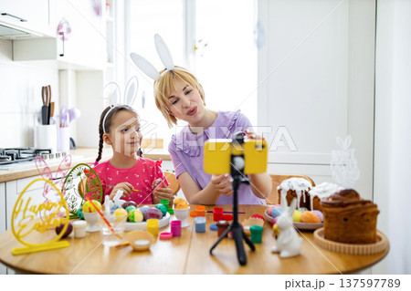 A mother and daughter wearing bunny ears are decorating Easter eggs and filming their activity on a smartphone A mother and daughter wearing bunny ears are decorating Easter eggs and filming their activity on a smartphone 137597789