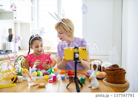 A mother and daughter wearing bunny ears paint Easter eggs together at a kitchen table while recording with a smartphone on a tripod A mother and daughter wearing bunny ears paint Easter eggs together at a kitchen table while recording with a smartphone on a tripod 137597791