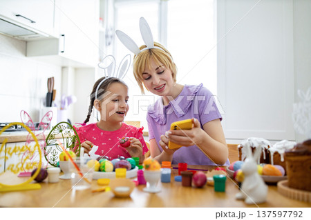 A mother and daughter wearing bunny ears are decorating Easter eggs together in a bright kitchen, looking at a smartphone for inspiration A mother and daughter wearing bunny ears are decorating Easter eggs together in a bright kitchen, looking at a smartphone for inspiration 137597792