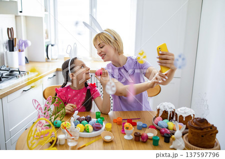 A mother and daughter wearing bunny ears are decorating Easter eggs in a bright kitchen, capturing the moment with a selfie A mother and daughter wearing bunny ears are decorating Easter eggs in a bright kitchen, capturing the moment with a selfie 137597796