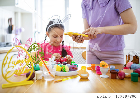 A mother photographs her daughter painting colorful Easter eggs at a kitchen table adorned with festive decorations and craft supplies 137597800