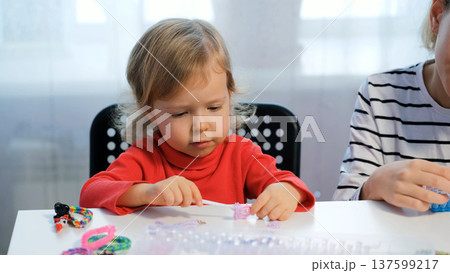 A cute girl makes a rubber band bracelet with her older sister while enjoying a creative activity at home A cute girl makes a rubber band bracelet with her older sister while enjoying a creative activity at home 137599217
