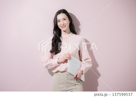 ピンクの背景でパソコンを持つかわいいオフィスカジュアルの日本人女性のカメラ目線の笑顔 137599537