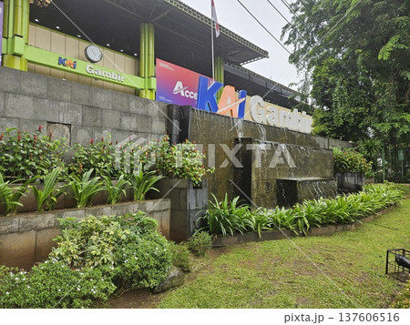 The iconic facade of Gambir Railway Station in Jakarta features the bold KAI Gambir signage above a tiered stone water fountain, surrounded by lush green garden plants under a gloomy overcast sky. The iconic facade of Gambir Railway Station in Jakarta features the bold KAI Gambir signage above a tiered stone water fountain, surrounded by lush green garden plants under a gloomy overcast sky. 137606516