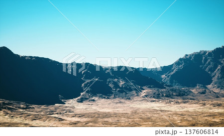 Sunlit desert valley under clear sky, wide panoramic view of rocky mountains and dry scrub, midday light casting deep shadows across rugged crater 137606814