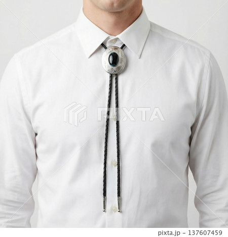 Man wearing a white shirt with a classic bolo tie featuring a silver clasp and black braided leather cord, isolated on a light background	 137607459