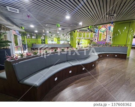 A modern waiting lounge featuring curved grey cushioned benches with integrated power outlets, decorated with festive green hanging ornaments and potted plants under a contemporary slatted ceiling. A modern waiting lounge featuring curved grey cushioned benches with integrated power outlets, decorated with festive green hanging ornaments and potted plants under a contemporary slatted ceiling. 137608362
