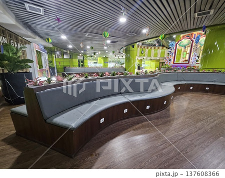 A modern waiting lounge featuring curved grey cushioned benches with integrated power outlets, decorated with festive green hanging ornaments and potted plants under a contemporary slatted ceiling. A modern waiting lounge featuring curved grey cushioned benches with integrated power outlets, decorated with festive green hanging ornaments and potted plants under a contemporary slatted ceiling. 137608366