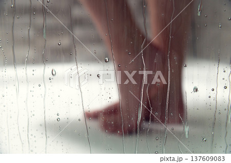 Female Bare feet of a person standing on a wet shower floor, with water droplets on a glass door creating a blurred effect in a modern bathroom setting 137609083