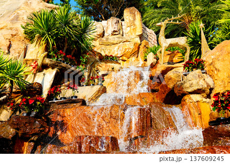 Waterfall flows down rocks surrounded by plants in sunny outdoor area in garden setting. Waterfall flows down rocks surrounded by plants in sunny outdoor area in garden setting. 137609245