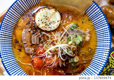 Lyon, france. December 29, 2023. Traditional japanese ramen noodle soup filled with tender pork, green onions, cherry tomatoes, and a lemon slice, overhead view Lyon, france. December 29, 2023. Traditional japanese ramen noodle soup filled with tender pork, green onions, cherry tomatoes, and a lemon slice, overhead view 137609874