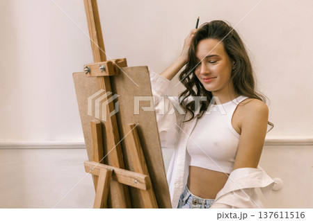 Young female artist standing near easel and holding pencil while working on painting in art studio, creative process and inspiration Young female artist standing near easel and holding pencil while working on painting in art studio, creative process and inspiration 137611516