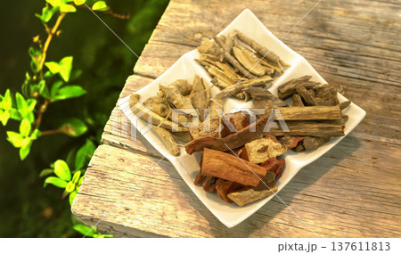 Assorted dried Thai medicinal herbs and wood chips on a white plate, placed on a rustic wooden table in a garden. Ideal for traditional medicine and natural healing themes 137611813