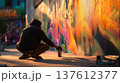 Street artist crouching beside colorful graffiti wall while spraying aerosol paint, urban street art scene with vibrant mural, creative expression and underground culture 137612377