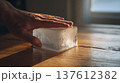 Hand touching melting ice cube on warm wooden surface, macro scene showing temperature contrast, water droplets and climate change concept with natural sunlight and physics experiment 137612382
