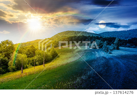 grassy rural fields in mountain landscape. day meet night. sun and moon over country road to forest through rolling hills. beautiful picture of a scenery on summer solstice weather with clouds on sky 137614276