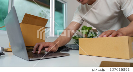 E-commerce Operations. Young man preparing packages while using laptop for online business. E-commerce Operations. Young man preparing packages while using laptop for online business. 137616891