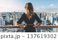 Woman with long hair stands on balcony overlooking city skyline with numerous high-rise buildings under a clear blue sky 137619302