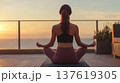 Woman practicing yoga in a meditative pose on a mat, with a sunset view over the ocean and plants in the background 137619305