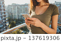 Woman in green shirt using smartphone on balcony with urban apartment buildings visible in the background during daylight 137619306