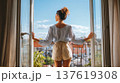 Woman in casual attire stands at open balcony doors, overlooking a cityscape with blue sky and clouds in the background 137619308