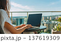 Woman using laptop on balcony with city skyline view, modern buildings and greenery visible in the background 137619309
