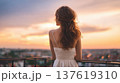 Faceless woman in white dress gazes at sunset from balcony overlooking cityscape with blurred lights and colorful sky 137619310