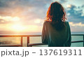 Faceless woman with wavy red hair gazes at sunset over ocean from balcony railing, clouds and warm colors in the sky 137619311