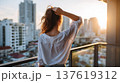 Woman with long hair in white shirt stands on balcony, overlooking city skyline at sunset with modern buildings in the background 137619312
