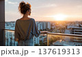 Woman with curly hair gazes at sunset from a balcony overlooking a city skyline with modern buildings and a clear sky 137619313