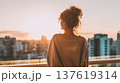 Faceless woman in gray sweater gazes at sunset over city skyline from balcony, urban buildings and soft light in the background 137619314