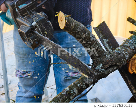 Person wearing jeans and a black sweater working outdoors, using a powerful electric chainsaw to cut tree branches into logs for firewood on a snowy winter day. Person wearing jeans and a black sweater working outdoors, using a powerful electric chainsaw to cut tree branches into logs for firewood on a snowy winter day. 137620115