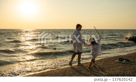 Two children in white bathrobes stroll along the sandy shore during golden hour. The child waves a stick, enjoying the warm sunset light, the gentle waves, and the carefree games on the seashore. Two children in white bathrobes stroll along the sandy shore during golden hour. The child waves a stick, enjoying the warm sunset light, the gentle waves, and the carefree games on the seashore. 137620142