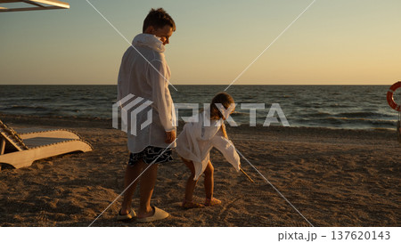 Two children, a boy and a girl, drawing on the sandy beach with a stick next to the sea during a warm evening golden hour, enjoying creativity and togetherness on their summer vacation. 137620143