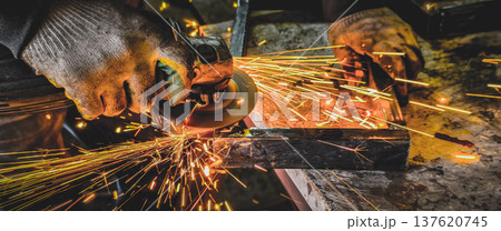 Worker hands wearing protective gloves operating an angle grinder, creating a shower of bright orange sparks while cutting or shaping metal in an industrial workshop. 137620745