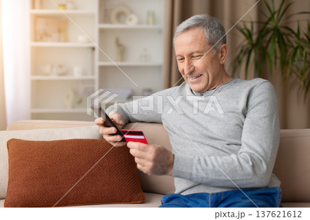 Senior man sitting on sofa using smartphone, holding credit card at home 137621612