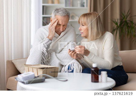 Senior woman checking thermometer while caring for sick husband wrapped in blanket 137621619