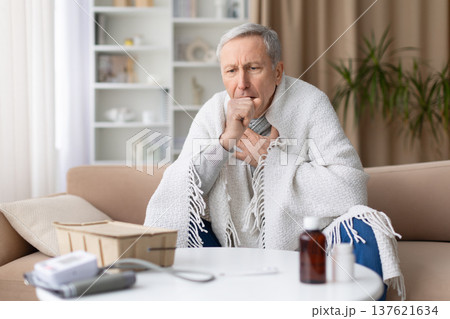 Senior man sitting on sofa wrapped in blanket while coughing at home Senior man sitting on sofa wrapped in blanket while coughing at home 137621634