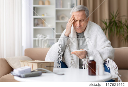 Senior man sitting on sofa at home checking thermometer while feeling unwell Senior man sitting on sofa at home checking thermometer while feeling unwell 137621662