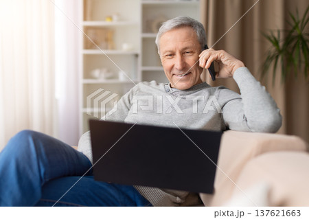 Smiling senior man relaxing on sofa, talking on smartphone and using laptop 137621663