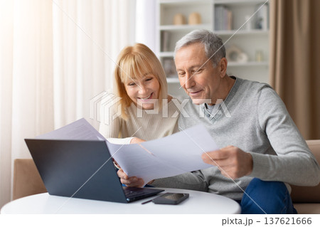 Senior couple reviewing documents while sitting at table with laptop at home Senior couple reviewing documents while sitting at table with laptop at home 137621666