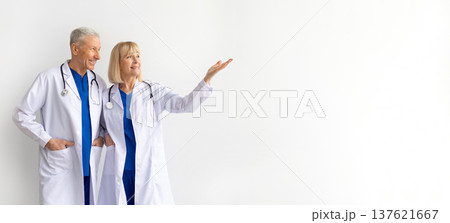 Two mature doctors in white coats standing on white background, pointing aside Two mature doctors in white coats standing on white background, pointing aside 137621667