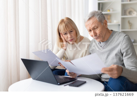 Concerned senior couple reading financial documents at home while sitting with laptop Concerned senior couple reading financial documents at home while sitting with laptop 137621669