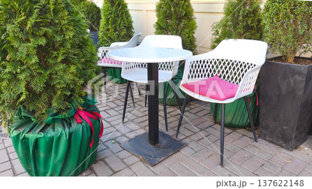 Outdoor cafe table with modern white chairs and pink cushions surrounded by green plants, creating a cozy terrace atmosphere for relaxation, conversation, and urban lifestyle. Outdoor cafe table with modern white chairs and pink cushions surrounded by green plants, creating a cozy terrace atmosphere for relaxation, conversation, and urban lifestyle. 137622148
