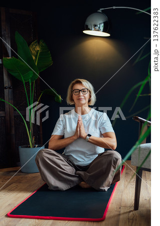 Middle aged woman practicing yoga, meditation at home, sitting in Namaste position on floor mat in modern Scandi dark interior home. Meditating, relaxed, eyes closed. Mental health and well being. Middle aged woman practicing yoga, meditation at home, sitting in Namaste position on floor mat in modern Scandi dark interior home. Meditating, relaxed, eyes closed. Mental health and well being. 137622383