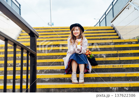 Portrait of young smiling woman with fresh tulips and coffee cup sitting on stairs at the city street enjoying the moment. Urban street fashion. Springtime yellow mood. Happiness and positive vibes Portrait of young smiling woman with fresh tulips and coffee cup sitting on stairs at the city street enjoying the moment. Urban street fashion. Springtime yellow mood. Happiness and positive vibes 137622386