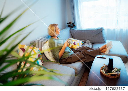 Middle aged woman relaxing with cup of hot drink on sofa in hygge interior home. Cozy comfortable winter or autumn weekends. Take a break and relax. Lady enjoy calm mood, well being alone Middle aged woman relaxing with cup of hot drink on sofa in hygge interior home. Cozy comfortable winter or autumn weekends. Take a break and relax. Lady enjoy calm mood, well being alone 137622387
