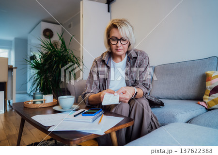 Middle aged woman reviewing expenses receipts while sitting on couch with coffee and phone in cozy living space. Managing personal finances with mobile app . Checking financial balance, budgeting Middle aged woman reviewing expenses receipts while sitting on couch with coffee and phone in cozy living space. Managing personal finances with mobile app . Checking financial balance, budgeting 137622388