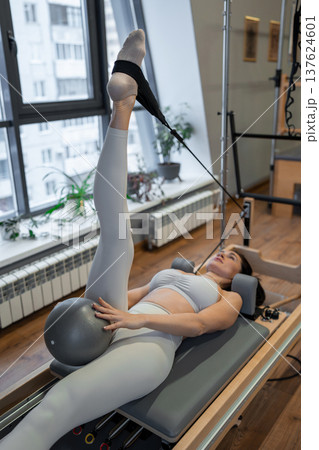 Woman in sportswear holding pilates pose on reformer machine with legs raised in bright studio. Pilates, legs raised, sportswear, reformer, stretching. Woman in sportswear holding pilates pose on reformer machine with legs raised in bright studio. Pilates, legs raised, sportswear, reformer, stretching. 137624601